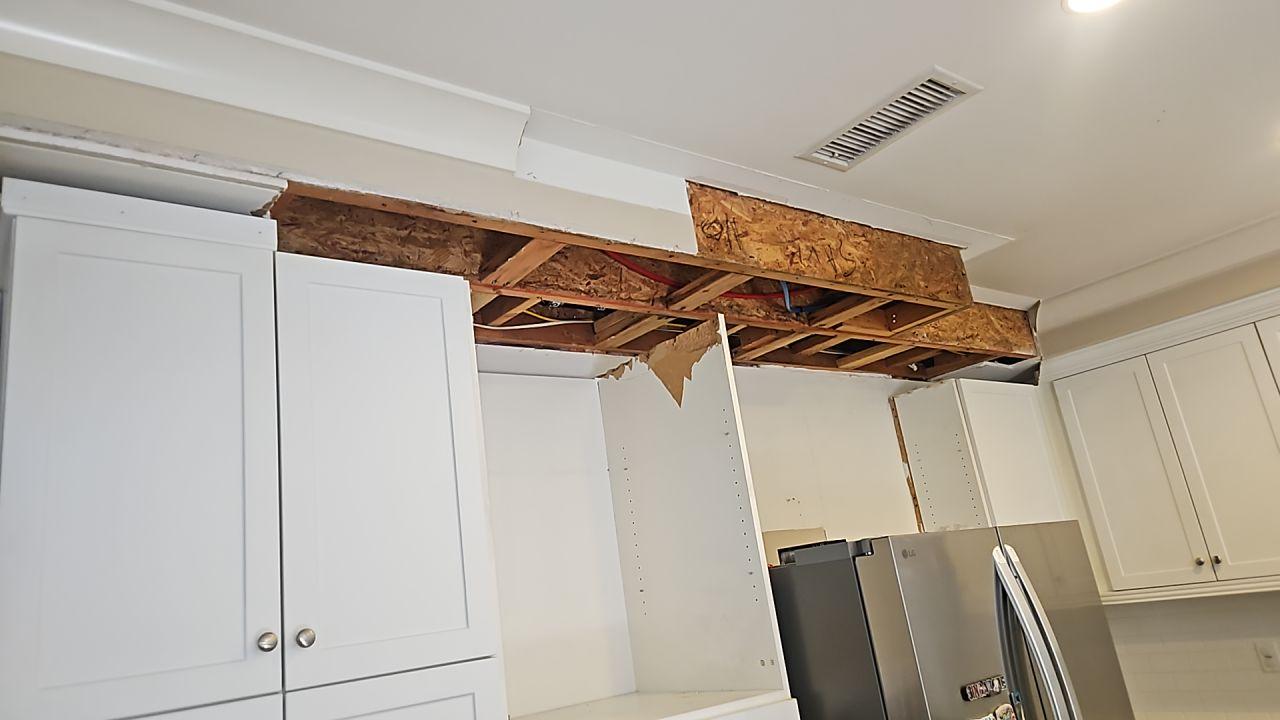 Water damage mitigation in the kitchen showing the ceiling area above cabinets exposed for structural drying and repair following a plumbing leak, revealing saturation and necessary water extraction access for restoration.